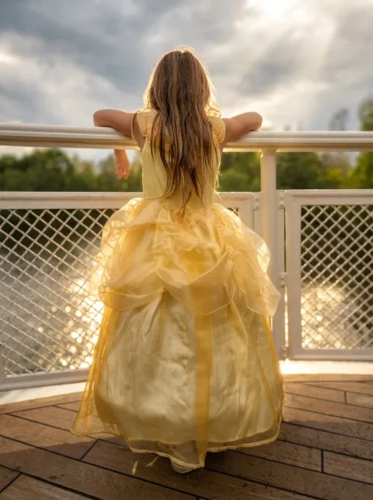 A girl in a yellow Belle dress, photographed from behind by a railing facing the water at Disneyland Paris